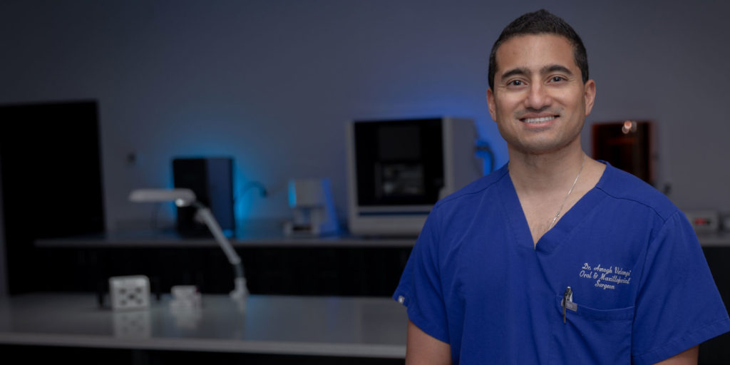 Oral and maxillofacial surgeon, Dr. Amogh Verangi in blue scrubs standing in Smile Now dental lab